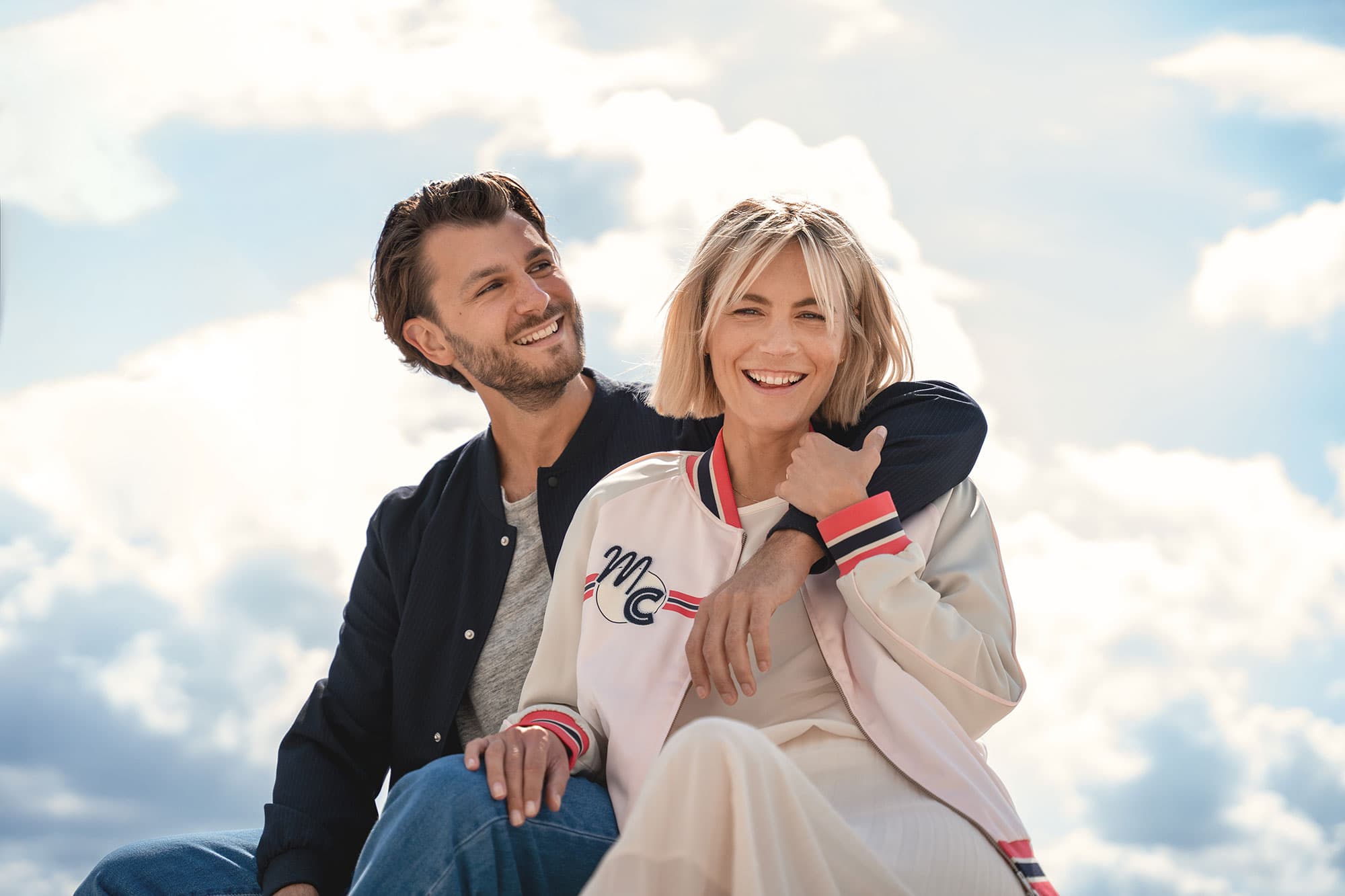 Ein lächelndes Paar sitzt im Freien vor dem Hintergrund eines hellen, bewölkten Himmels. Die Frau trägt eine helle Jacke, der Mann eine dunkle Jacke.