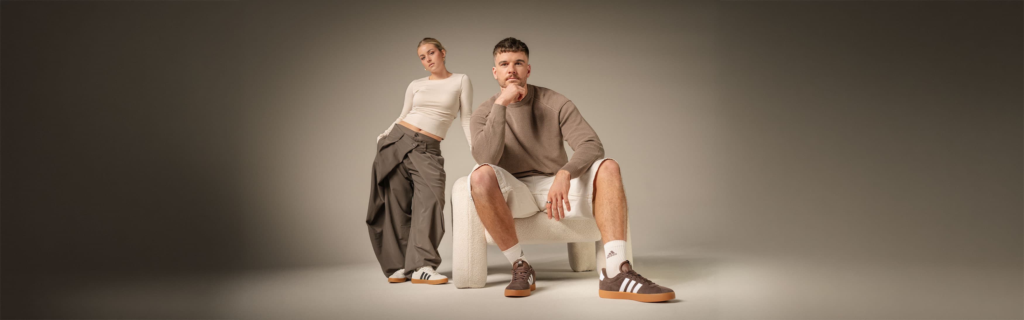 Zwei Personen in Freizeitkleidung und Adidas-Turnschuhen posieren in einer minimalistischen Umgebung, eine sitzt, die andere steht.